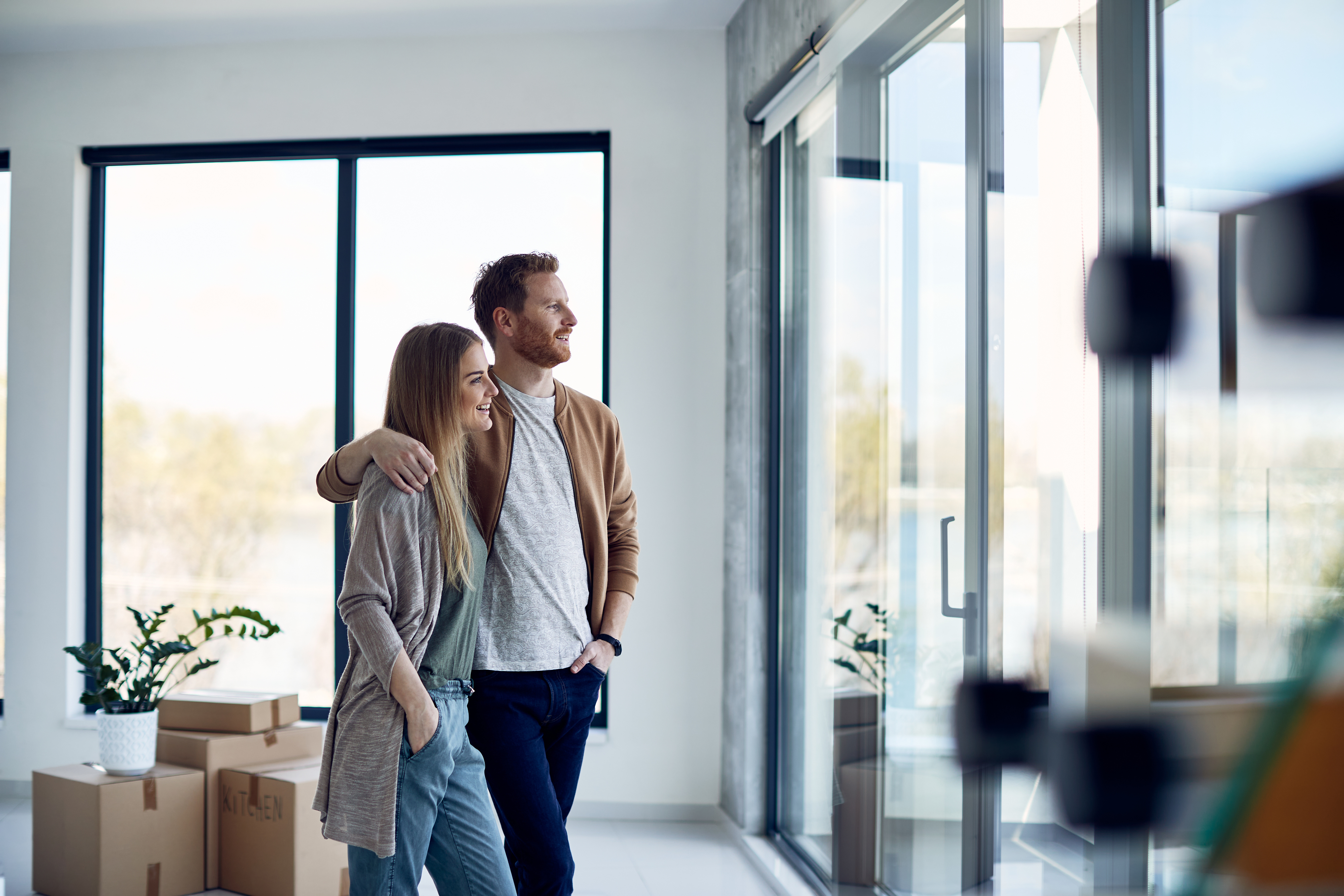Titelbild Startseite Junges glückliches Paar genießt den Ausblick durch das Fenster in ihrem neuen Zuhause.